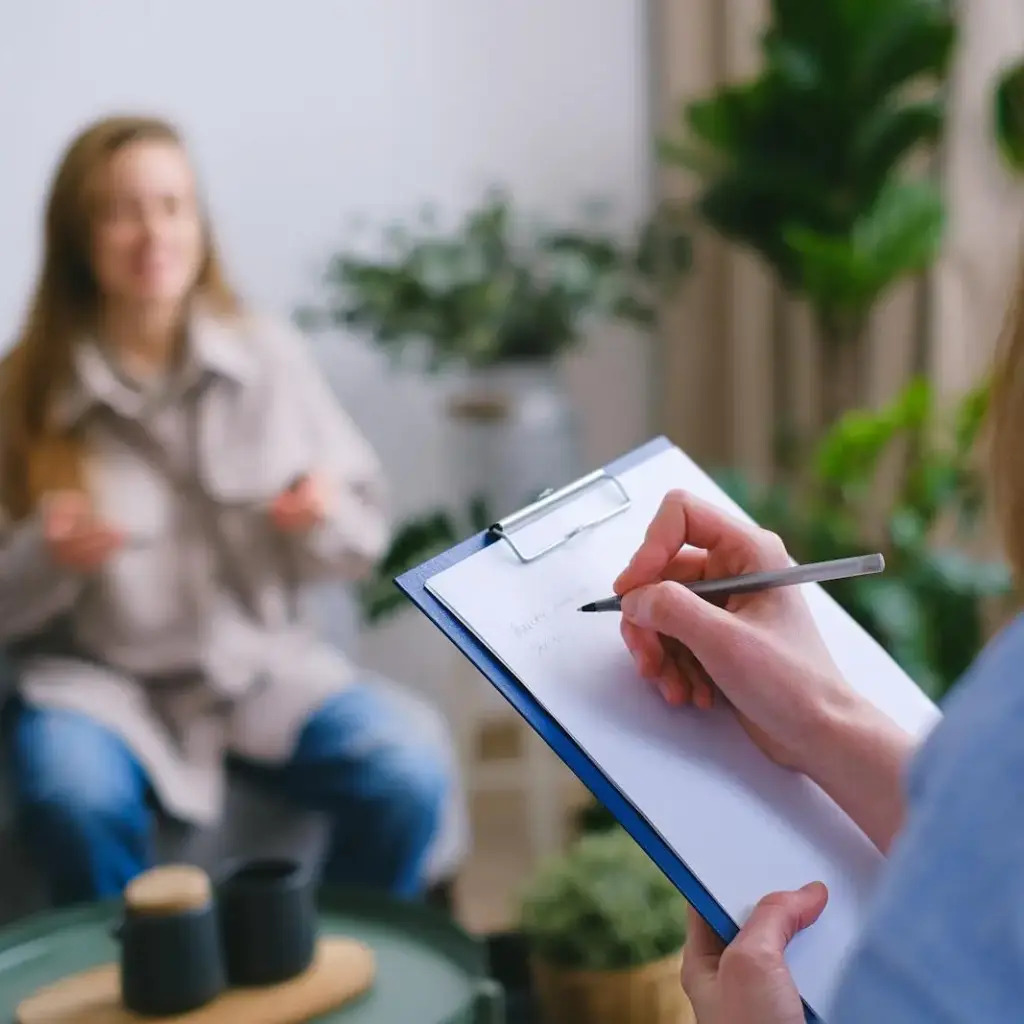 Unrecognizable professional female psychologist writing on clipboard while sitting against client on blurred background during psychotherapy session in light office